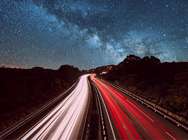 一条高速公路的长时间曝光夜景照片，在晴朗、繁星点点的天空和银河下，有红色和白色的光迹。