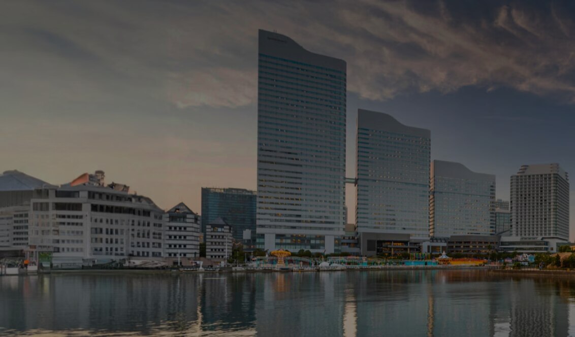 Waterfront cityscape at sunset with modern high-rise buildings reflected in the calm water, a large Ferris wheel on the right, and a tree-lined promenade with benches in the foreground.