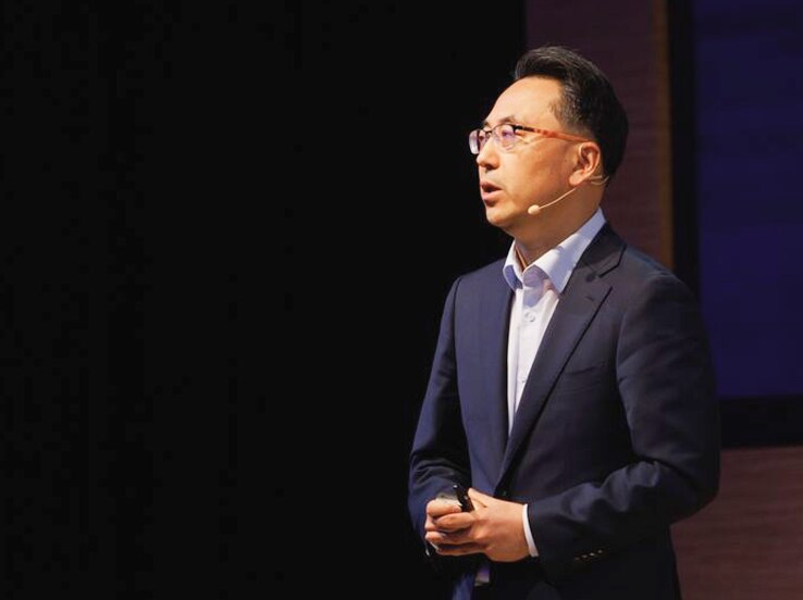 A keynote speaker in a navy suit delivers a professional presentation on stage at a Samsung technology event.