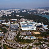 鸟瞰一个大型半导体设施综合体，周围是道路、绿地和远处的城市发展。
