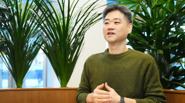 安全与电力产品开发团队的任务负责人姜贤宰，身穿绿色毛衣，在布满植物的办公室里以轻松的表情说话